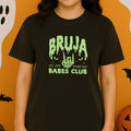 Woman wearing a black t-shirt with 'BRUJA BABES CLUB' text in a Halloween-themed setting with pumpkins and ghosts.