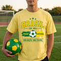 Person wearing a yellow 'Brásíl' t-shirt holding a soccer ball on a sports field.