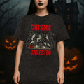 Woman wearing a dark t-shirt with graphic design cafecito and chisme and text in a Halloween-themed setting with pumpkins and a haunted house.