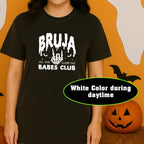 Person wearing a black t-shirt with 'BRUJA BABES CLUB' text in a Halloween-themed setting with pumpkins and ghosts.