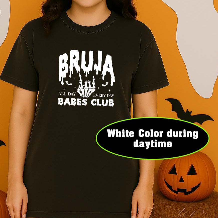 Person wearing a black t-shirt with 'BRUJA BABES CLUB' text in a Halloween-themed setting with pumpkins and ghosts.