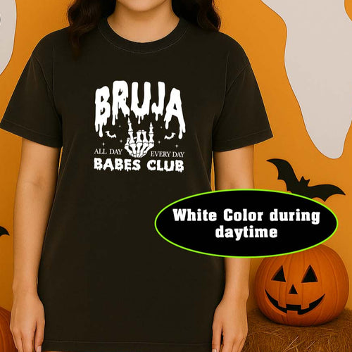 Person wearing a black t-shirt with 'BRUJA BABES CLUB' text in a Halloween-themed setting with pumpkins and ghosts.