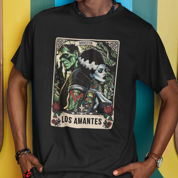 Person wearing a black t-shirt with 'Los Amantes' design and a cap, standing against a colorful wall.