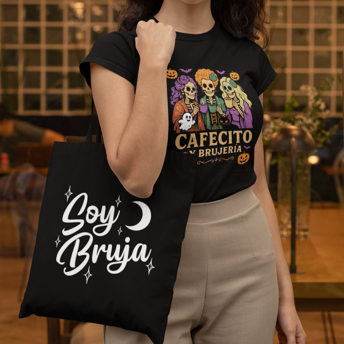 Woman wearing a black t-shirt with a graphic design and holding a black tote bag with 'Soy Bruja' text.