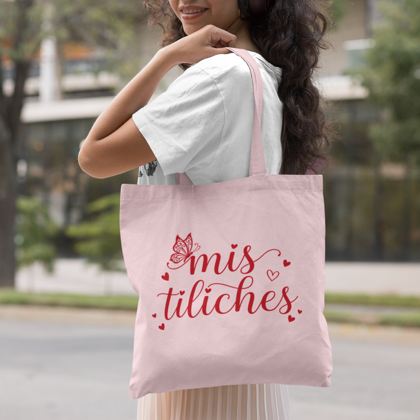 Woman holding a pink tote bag with 'mis tiliches' text, wearing headphones outdoors.